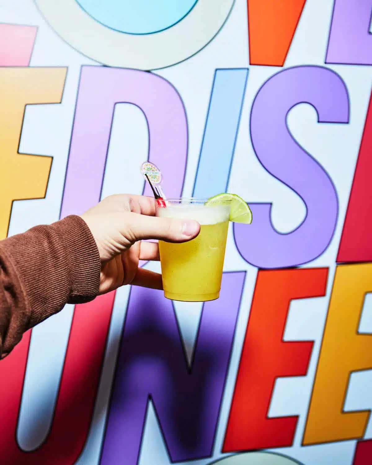 Hand holding a vibrant yellow cocktail with a lime wedge in front of a colorful, playful background featuring bold, oversized letters spelling "FESTIVAL FUN."