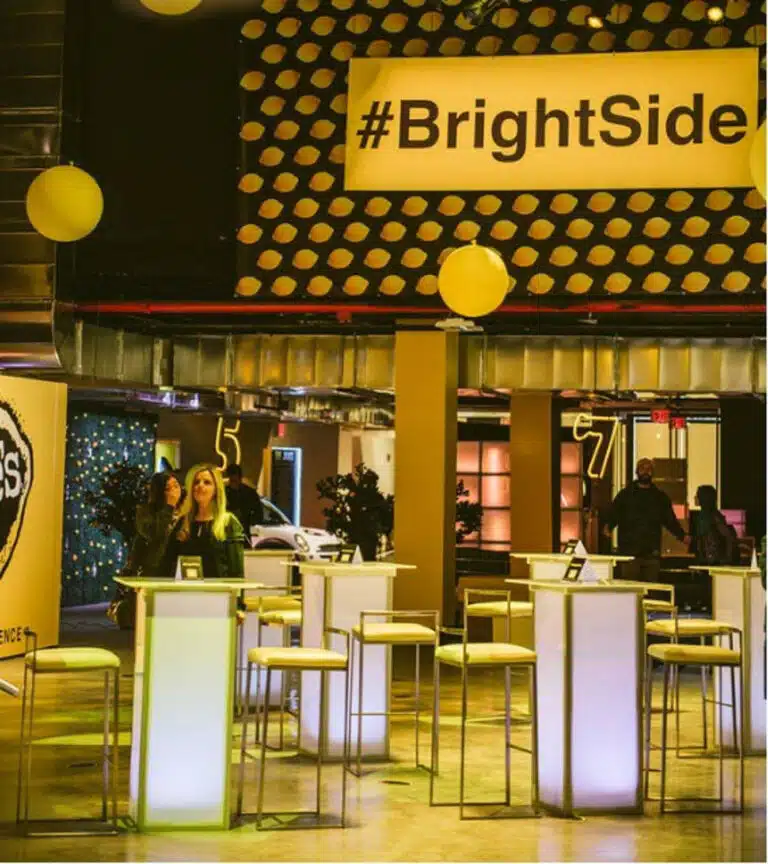Event space featuring illuminated high tables and stools, decorated with a #BrightSide banner and lemon-themed design, promoting social interaction and networking at events.