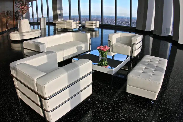 Modern lounge furniture setup featuring white sofas and armchairs, a glass coffee table with a floral arrangement, in a spacious, elegantly designed event space with panoramic city views.